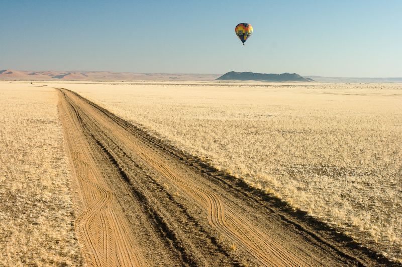 A small hot air balloon in the sky above a straight gravel track across the plain