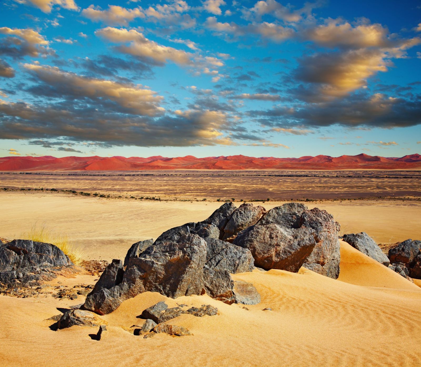Weathered boulders in the sand with red dunes and a cloudy sky behind them